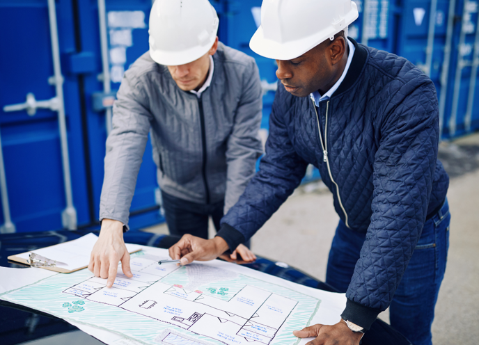 To ingeniører med sikkerhedshjelme på, der står og diskuterer en plan sammen udenfor på deres arbejdsplads